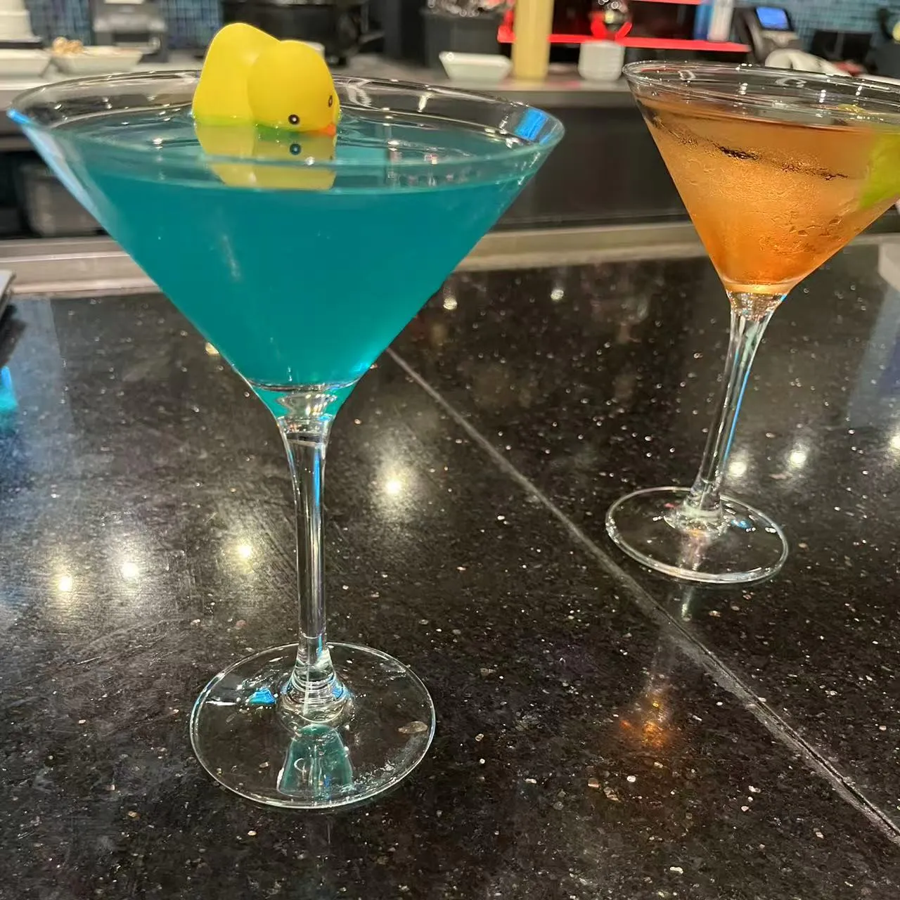 Bright blue martini cocktail topped with yellow rubber duck beside amber cocktail